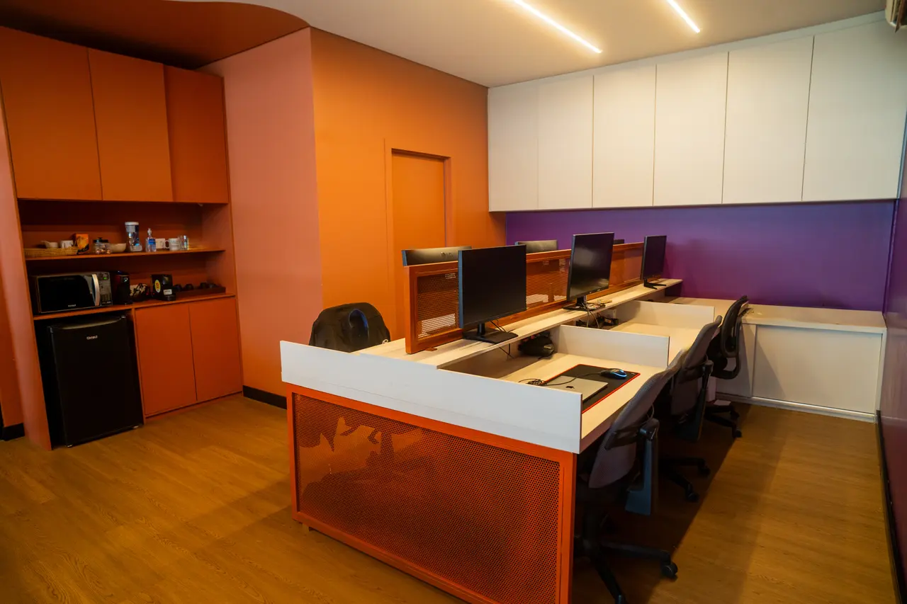 Bancada de trabalho com monitores e, ao lado, a copa com micro-ondas e frigobar.