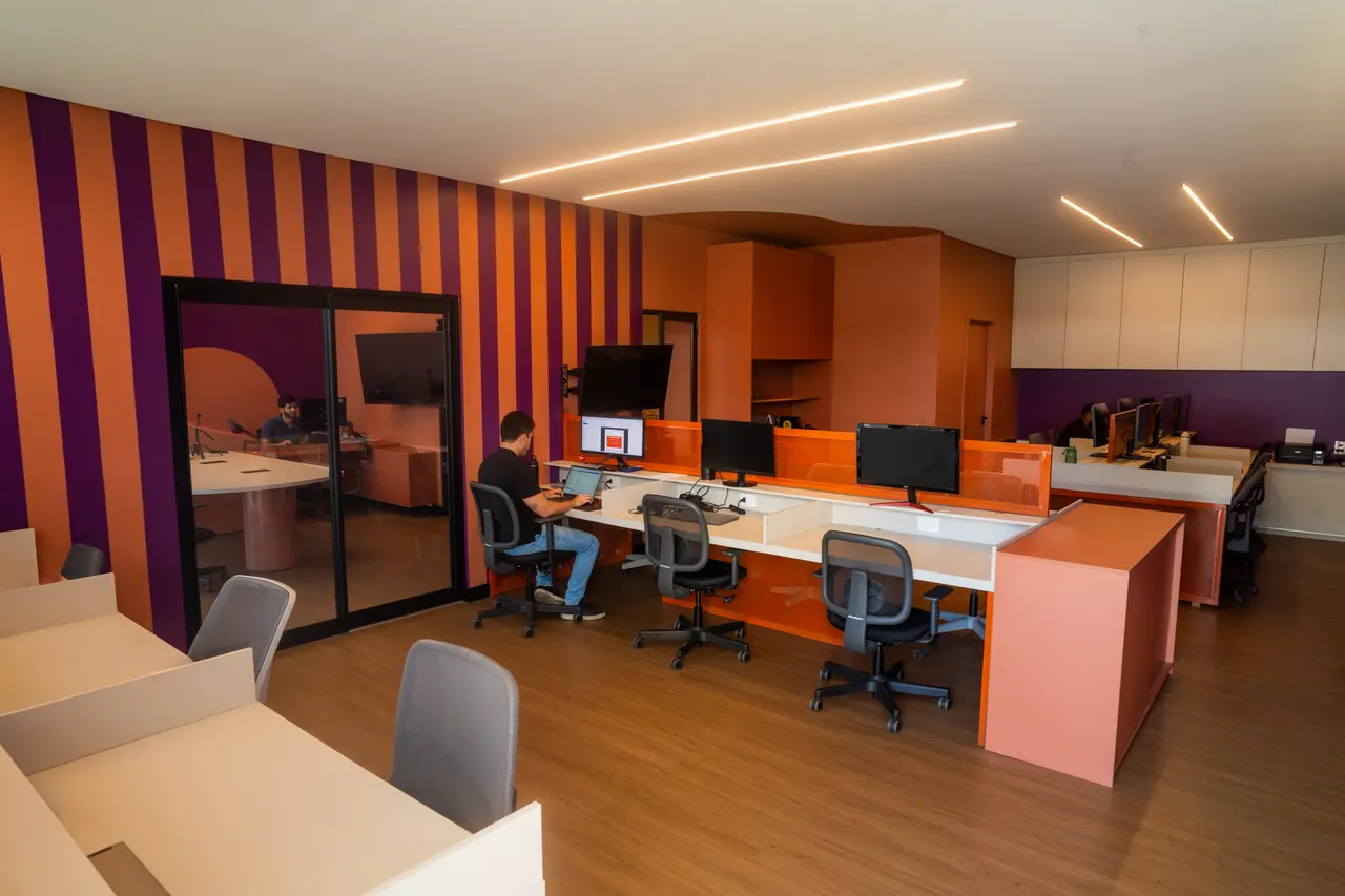 Sala da equipe com parede listrada em roxo e laranja, mesas com monitores e a sala de reunião envidraçada à esquerda.
